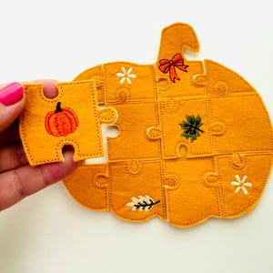 May include: A felt pumpkin puzzle with a missing piece held in a hand. The puzzle is orange with embroidered details including a pumpkin, leaves, and a bow. The missing piece has an embroidered pumpkin.