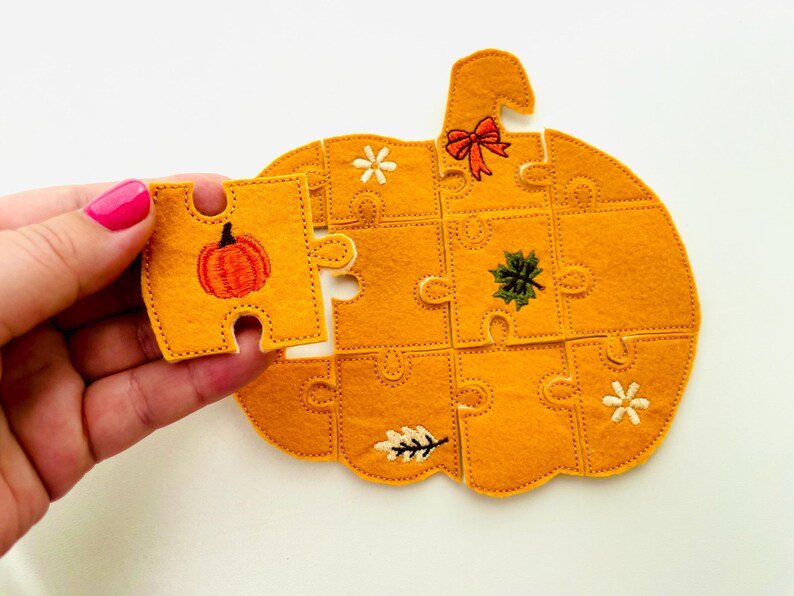 May include: A felt pumpkin puzzle with a missing piece held in a hand. The puzzle is orange with embroidered details including a pumpkin, leaves, and a bow. The missing piece has an embroidered pumpkin.
