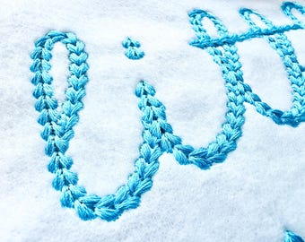 May include: A close-up of the word "with" embroidered in light blue thread on a white fabric background.