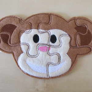 May include: A brown felt monkey puzzle piece with a white face, black eyes, a pink nose, and a white smile.
