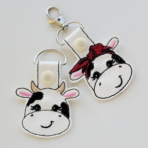 May include: Two white cow keychains with black spots. One cow has a red bandana. Both cows have black eyes and a black nose. The keychains are made of white leather and have a silver metal key ring.