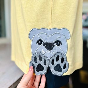 以下が含まれることがあります： 黄色のTシャツに、グレーの刺繍でブルドッグのデザインが施されています。ブルドッグはシャツの後ろから顔を出しており、足がのぞいています。