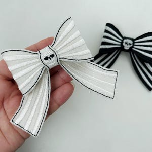 May include: Two embroidered bow appliques. One is white with black trim and a skull design, the other is black and white striped with a skull. These bows are a fun accessory for clothing or crafts.