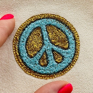 Könnte beinhalten: Ein gestickter Peace-Zeichen-Patch in Blau und Gold. Der Patch ist rund und hat einen goldenen Rand.