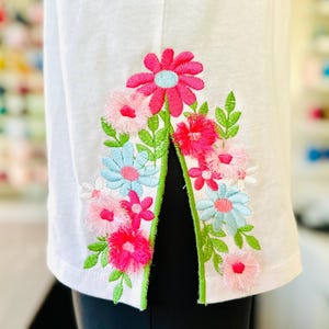 May include: White t-shirt with a floral embroidery design on the sleeve. The embroidery features pink, blue, and green flowers.