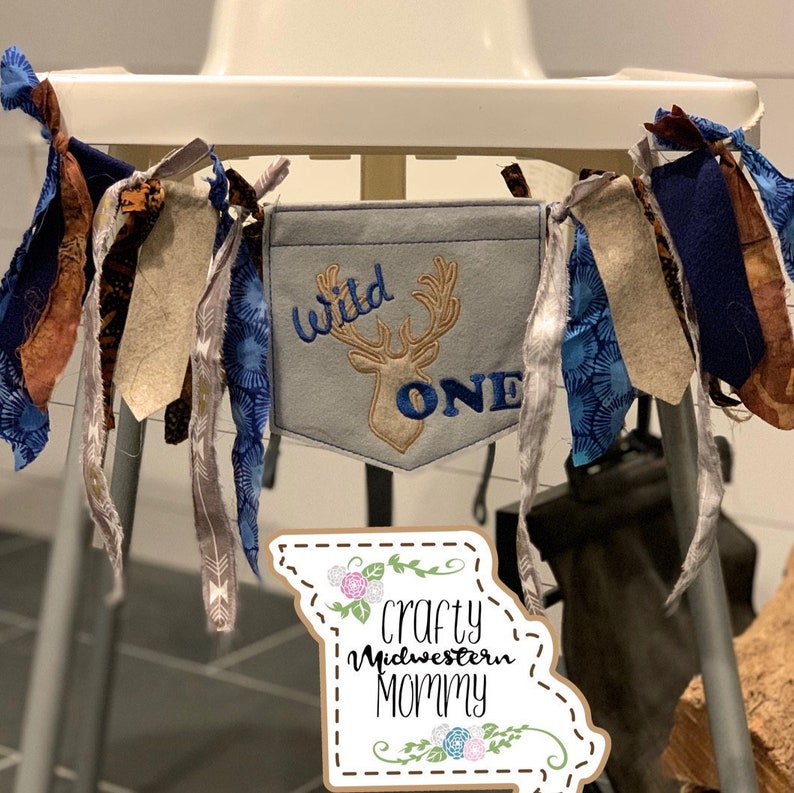 Op de afbeelding: Een blauwe en bruine stoffen banner met de tekst "Wild One" en een hert silhouet. De banner hangt aan een hoge stoel. Een bruin papieren bord met de tekst "Crafty Midwestern Mommy" is op de voorgrond.