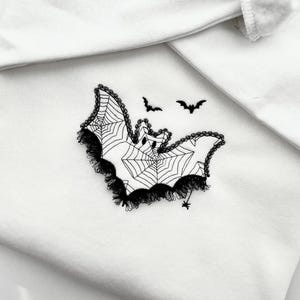 Puede incluir: Tela blanca con un diseño de murciélago bordado en negro. El murciélago tiene un patrón de telaraña y un ribete de encaje. Dos pequeñas siluetas de murciélago están encima del murciélago. Una pequeña araña está debajo.