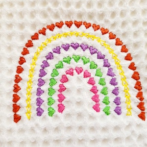 May include: A white waffle weave fabric with a rainbow embroidered in a series of hearts. The rainbow is made up of red, orange, yellow, green, purple, and pink hearts.