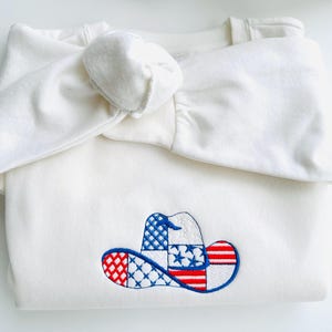 Könnte beinhalten: Ein gefaltetes, cremefarbenes Sweatshirt mit passendem geknotetem Stirnband. Das Sweatshirt hat ein gesticktes Cowboyhut-Design in Rot, Weiß und Blau, das der amerikanischen Flagge ähnelt. Der Hut ist blau umrandet.