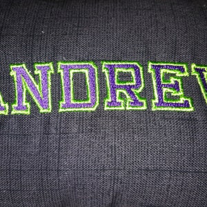 May include: A black fabric pillow with the name "ANDREW" embroidered in green and purple thread.