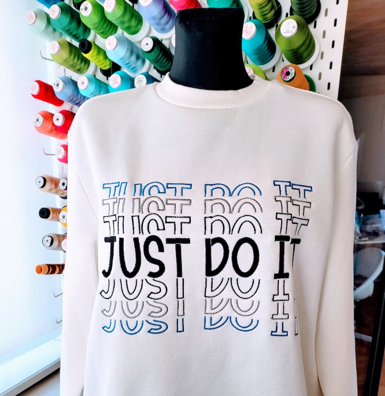 May include: White sweatshirt with a black embroidered "JUST DO IT" graphic. The text is repeated multiple times in a stacked, layered design with blue and gray outlines.