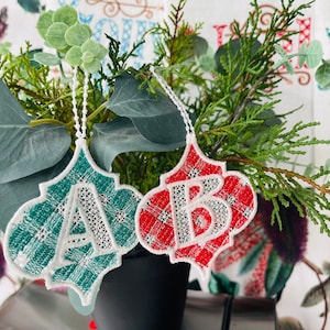 May include: Two Christmas ornaments with a white lace design and the letters A and B. The ornament with the letter A is green and the ornament with the letter B is red.
