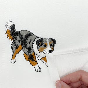 May include: A white kitchen towel featuring an embroidered Australian Shepherd dog. The dog has a tri-color coat of gray, white, and tan, with a fluffy tail and a focused expression. The fabric of the towel has a subtle herringbone weave pattern.