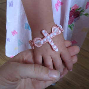 May include: A pink and white cross-shaped bracelet with a white centre. The bracelet is on a child's wrist.