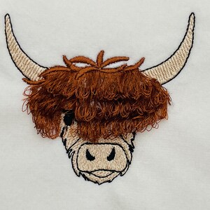 May include: Embroidered design of a Highland cow head with brown fur and white horns. The cow's face is white with black eyes and a black nose.