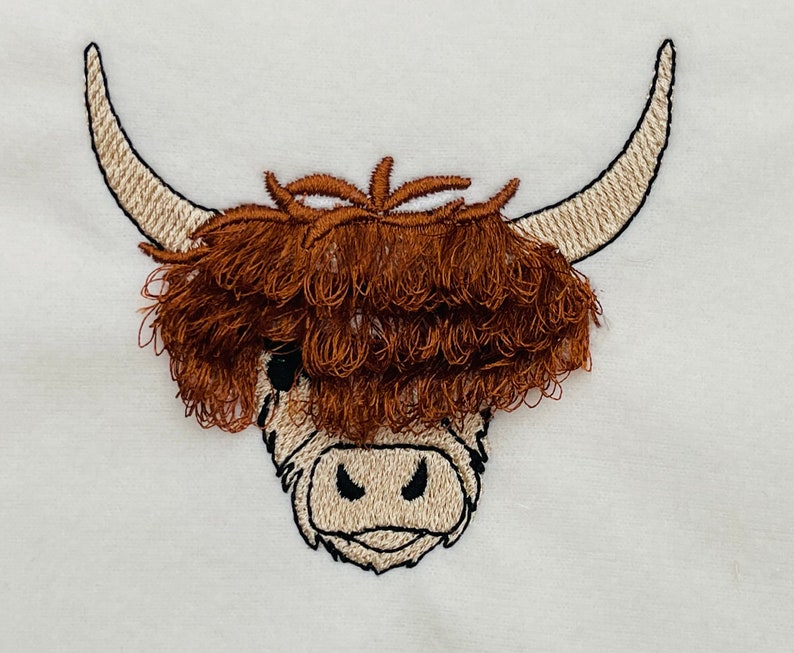 May include: Embroidered design of a Highland cow head with brown fur and white horns. The cow's face is white with black eyes and a black nose.