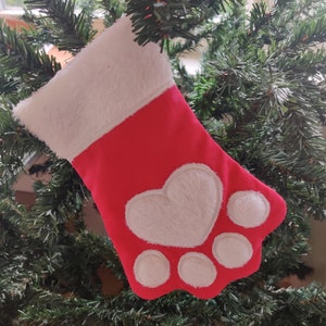 May include: Red Christmas stocking with a white paw print design. The paw print has a heart in the center.