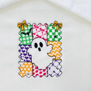 May include: White fabric with an embroidered Halloween design. The design features a white ghost in the center, surrounded by a patchwork border of colorful squares with various patterns and small yellow bows.