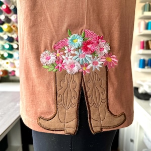 May include: A brown t-shirt with an embroidered design of a pair of cowboy boots with a bouquet of pink, white, and blue flowers.
