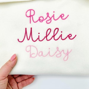 Puede incluir: Una tela blanca con tres nombres bordados en tonos rosas: "Rosie", "Millie" y "Daisy". Los nombres están bordados en cursiva, con diferentes tonos de rosa. La tela está sujeta por una mano con esmalte de uñas rosa.