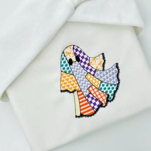 May include: A white fabric item featuring an embroidered ghost design. The ghost is composed of colorful patchwork with orange, purple, and teal accents, outlined in black lace. The item is folded, with a white fabric edge visible.