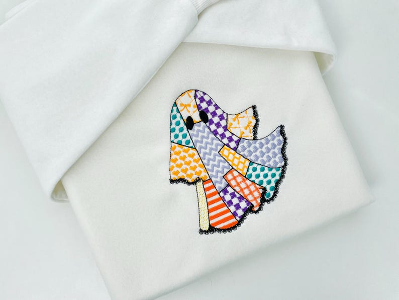 May include: A white fabric item featuring an embroidered ghost design. The ghost is composed of colorful patchwork with orange, purple, and teal accents, outlined in black lace. The item is folded, with a white fabric edge visible.