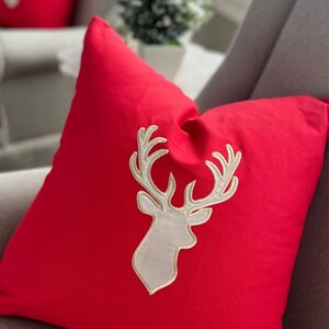 Op de afbeelding: Een rood kussen met een wit geborduurd rendiermotief. Het rendier heeft gouden geweien.