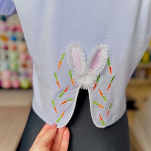 May include: White t-shirt with a white bunny rabbit applique on the back. The bunny has pink ears and is surrounded by embroidered green and orange carrots.