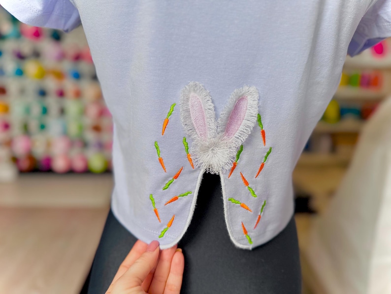 May include: White t-shirt with a white bunny rabbit applique on the back. The bunny has pink ears and is surrounded by embroidered green and orange carrots.