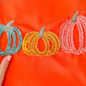 May include: Three embroidered pumpkins on an orange background. The pumpkins are teal, yellow, and pink.