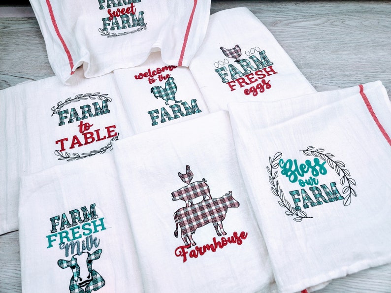 May include: Set of six white kitchen towels with red and green embroidered designs. The towels feature various farm-themed phrases, including "Farm to Table", "Welcome to the Farm", "Farm Fresh Eggs", "Farm Fresh Milk", "Bless Our Farm", and "Sweet Farm".