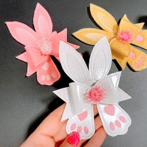 May include: Three felt bunny hair bows with pink, white, and yellow felt. Each bow has a small pink felt paw print on the bottom.