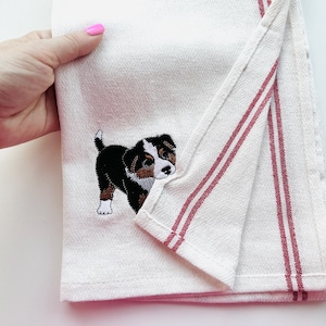 May include: White kitchen towel with a red striped border and an embroidered puppy design. The puppy is black, brown, and white. The towel is folded and held by a hand with pink nail polish.