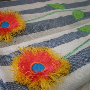 May include: Two embroidered flowers on a white and blue striped fabric. The flowers are yellow and red with a blue center.