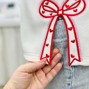 Op de afbeelding: Wit sweatshirt met een groot rood strikdesign. De strik is versierd met kleine rode hartjes en een roze kanten rand. Het ontwerp is geborduurd op de voorkant van het sweatshirt.