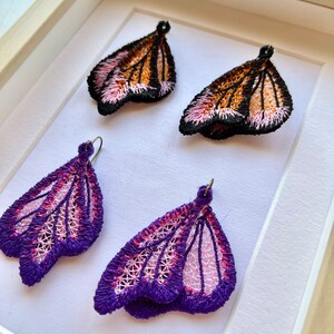 May include: A pair of butterfly-shaped earrings with pink and purple embroidery. The earrings are displayed in a white frame.