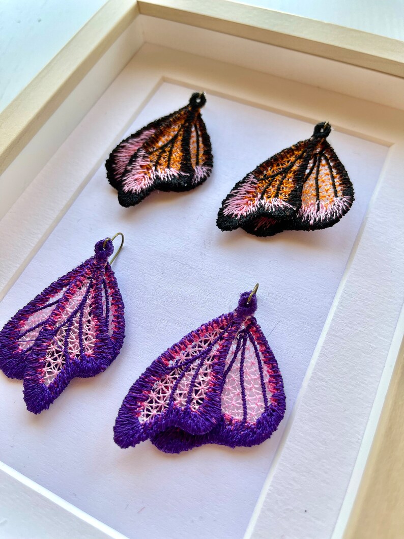 May include: A pair of butterfly-shaped earrings with pink and purple embroidery. The earrings are displayed in a white frame.