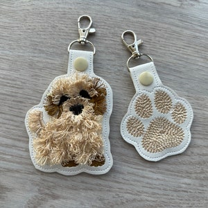 Op de afbeelding: Twee witte leren sleutelhangers met zilveren sleutelringen. De ene sleutelhanger heeft een geborduurde bruine en witte hond, de andere heeft een geborduurde bruine poot.