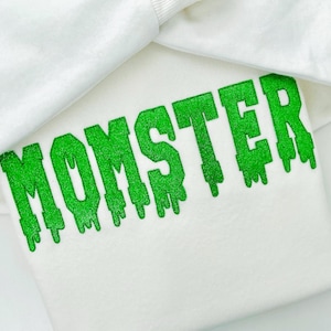May include: A white sweatshirt with the word "MOMSTER" in dripping green glitter lettering. The sweatshirt is folded, with the top portion of the garment visible, creating a layered effect.
