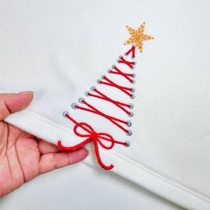Puede incluir: Sudadera blanca con un diseño de árbol de Navidad bordado en rojo. El árbol está creado con hilo rojo entrelazado a través de ojales plateados, coronado con una estrella dorada. Una cinta roja está en la base del árbol.