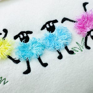 以下が含まれることがあります： 白い生地に刺繍された羊。羊は黒い顔と足を持ち、ふわふわの黄色、青、ピンクのウールが付いています。緑の刺繍された草が下にあります。羊は遊び心のあるポーズをとっています。