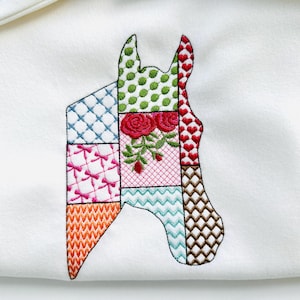 May include: A white fabric with an embroidered horse head design. The horse head is made up of different colored fabric squares with various patterns, including polka dots, hearts, and zigzags. There are two red roses embroidered on the horse's head.