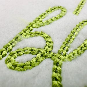 Puede incluir: Primer plano de texto bordado en color verde claro sobre tela blanca. La costura es gruesa y texturizada, creando un efecto en relieve. Las letras son estilizadas y cursivas, con apariencia hecha a mano. La estética general es suave y delicada.