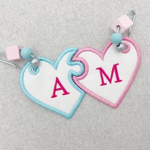 May include: Two heart-shaped fabric tags with puzzle-piece edges, featuring the embroidered letters A and M in pink. Each tag has a metal hook, a light pink wooden cube, and light blue and silver beads attached to the top corner.