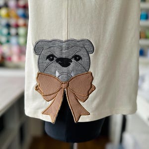 Könnte beinhalten: Ein weißer Stoff-Patch mit einem gestickten grauen Bulldoggen, der eine braune Schleife trägt.
