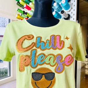 May include: A light green t-shirt with the words "Chill Please" embroidered in rainbow colors with a gold glitter finish. The shirt also features a gold glitter smiley face with sunglasses.