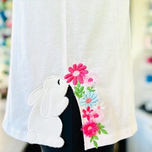 Può includere: Coniglio bianco ricamato con fiori rosa, blu e bianchi su una maglietta bianca.