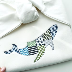 May include: White baby blanket with an embroidered whale design. The whale is composed of various patterned patches in shades of blue and green. The blanket is folded and tied at the top.
