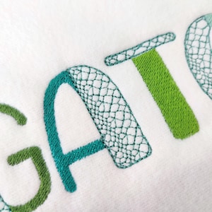 May include: Close-up of white fabric embroidered with the word "GATOR". The letters are in shades of green and teal. The letters "G" and "A" have a mesh-like pattern, while the "T" is solid green.