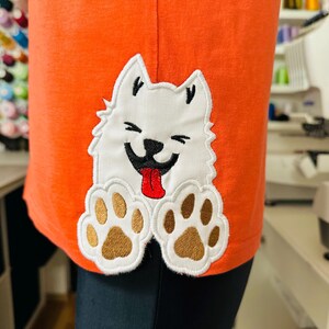 以下が含まれることがあります： オレンジ色のTシャツに、白い笑顔の犬のアップリケ。犬は黒い鼻と目、赤い舌、茶色の足跡があります。シャツは柔らかい素材で作られています。